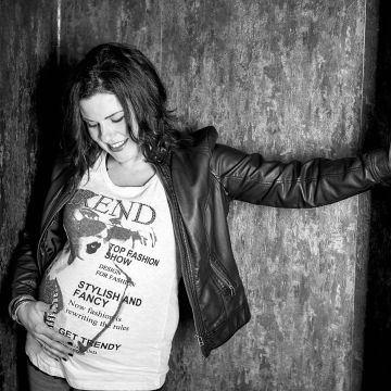 Schwangere in Jeans und T-Shirt lehnt an der Wand und schaut auf ihren schwangeren Bauch